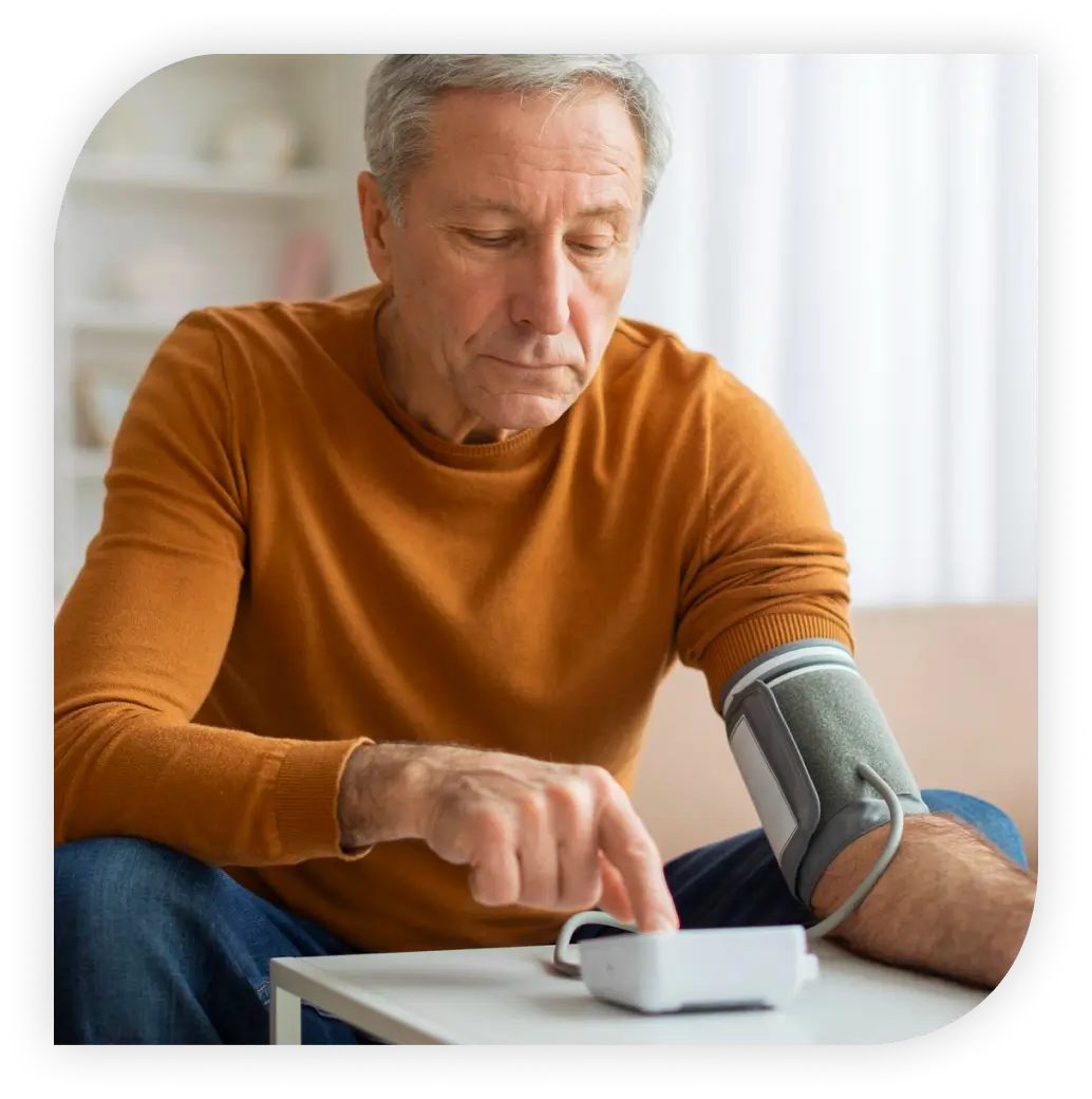 Un homme plus âgé, concentré, vêtu d'un pull orange, est assis et utilise un tensiomètre automatique sur son bras gauche, appuyant sur un bouton de la machine avec sa main droite.